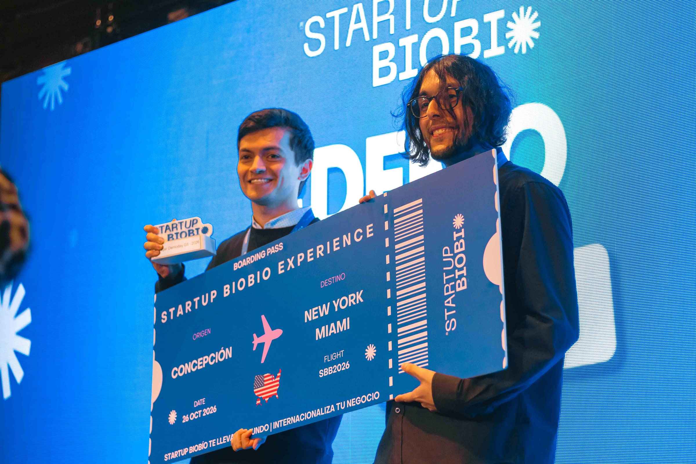 Dos integrantes de PAIR en el escenario de la final de Startup Biobío sosteniendo el pasaje simbólico del premio, un vuelo Concepción a Nueva York y Miami.